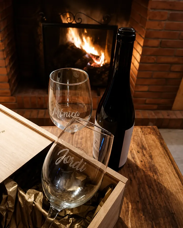 Dos copas grabadas con los nombres Mónica y Jordi, botella de vino y estuche de madera junto a la chimenea